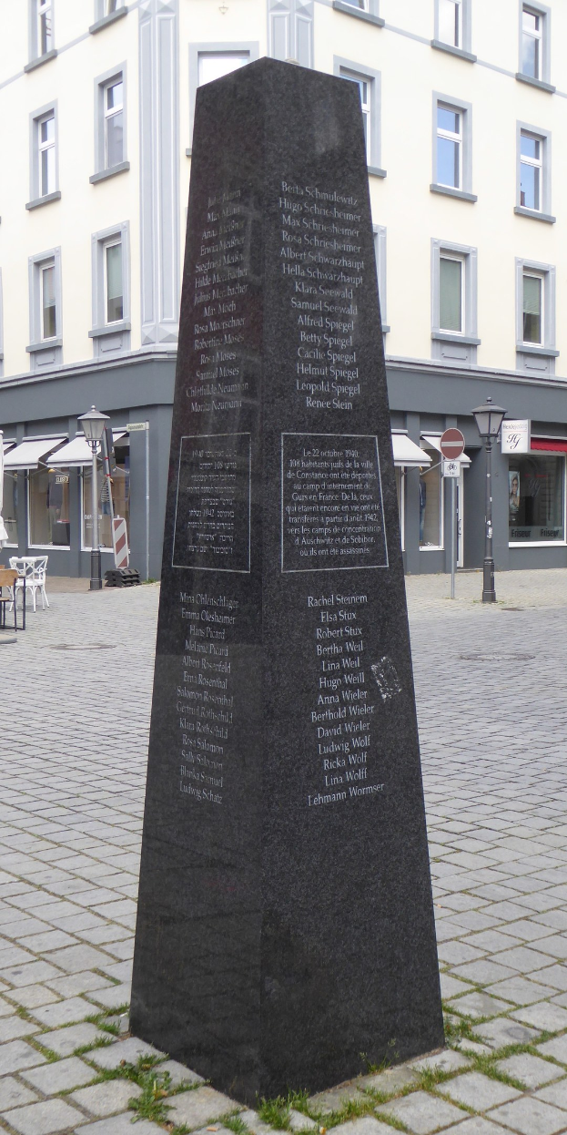 Ein hohes Denkmal mit eingravierter Schrift steht auf einem Platz, umgeben von Gebäuden, Straßenmöbeln und Menschen, mit dem Himmel im Hintergrund.