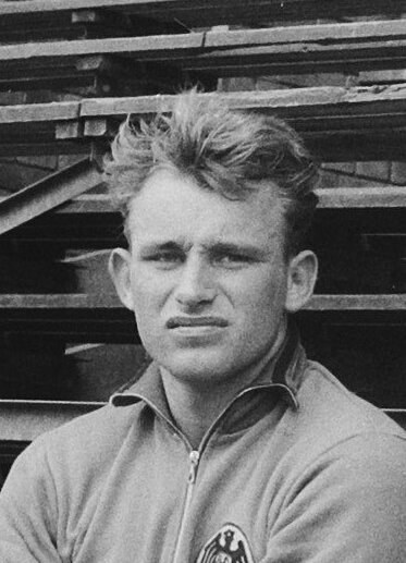 Black and white photo of a man in a jacket standing in front of stacked wooden planks, believed to be a legendary German footballer.