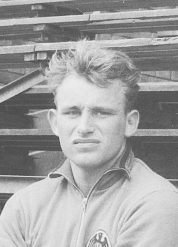 Black and white photo of a man in a jacket standing in front of stacked wooden planks, believed to be a legendary German footballer.