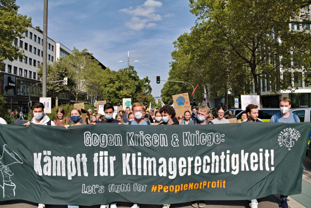 Eine Gruppe maskierter Personen hält ein Transparent mit der Aufschrift "Lass uns für die Menschen kämpfen, nicht für den Profit" vor einer Straßenszene mit Fahrzeugen, Bäumen, Gebäuden, Laternen und Ampeln unter einem bewölkten Himmel.