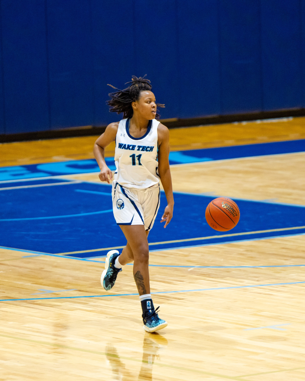 Frau in blauer und weißer Uniform, die einen Basketball auf einem Platz dribbelt, trägt ein weißes T-Shirt mit der Aufschrift "Wake Tech Women's Basketball" und blaue Schuhe, mit einer blauen Wand im Hintergrund.