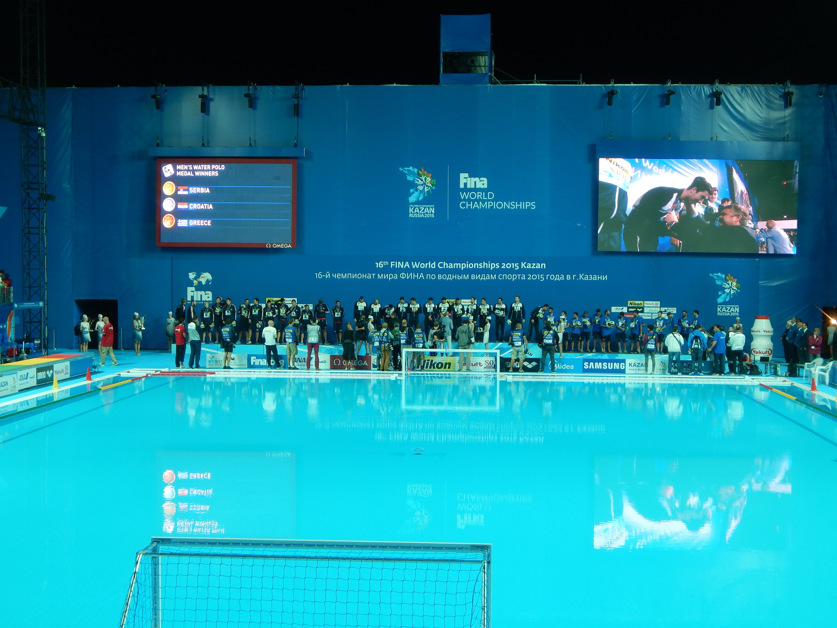 Gruppe von Menschen, die um ein Hallenbad mit Netz am Boden stehen, mit einem Wandbildschirm im Hintergrund, der "FINA World Championships 2015 Kazan" anzeigt.