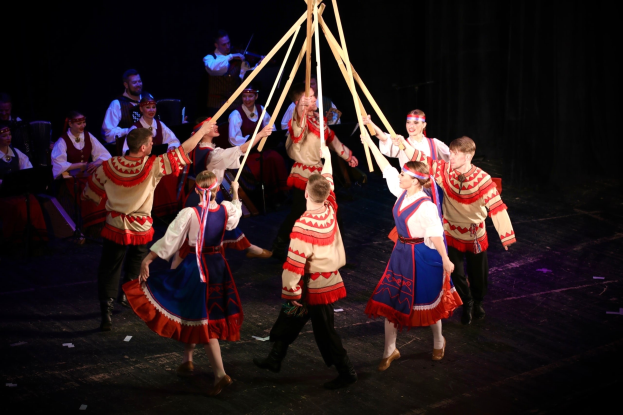 Eine Gruppe von Menschen in farbenfrohen Kostümen tanzt um einen Maibaum, hält Holzstäbe in den Händen, während Musiker im Hintergrund Instrumente spielen.
