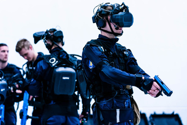 Eine Gruppe von Polizisten in schwarzen Uniformen und VR-Brillen steht zusammen, einer hält eine Waffe, mit verschiedenen Gegenständen im Hintergrund.