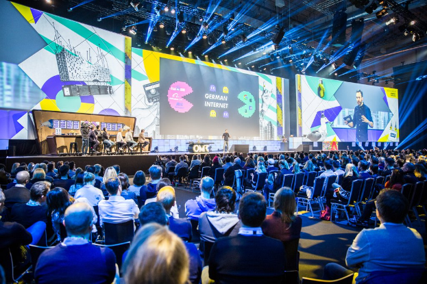 Eine große Menschenmenge, die auf Stühlen vor einer Bühne sitzt, mit einigen Individuen, die auf dem Podium in der foreground stehen, einem Tisch mit verschiedenen Gegenständen links und Bildschirmen und Lichtern im Hintergrund auf der Deutschen Internet-Konferenz 2019.