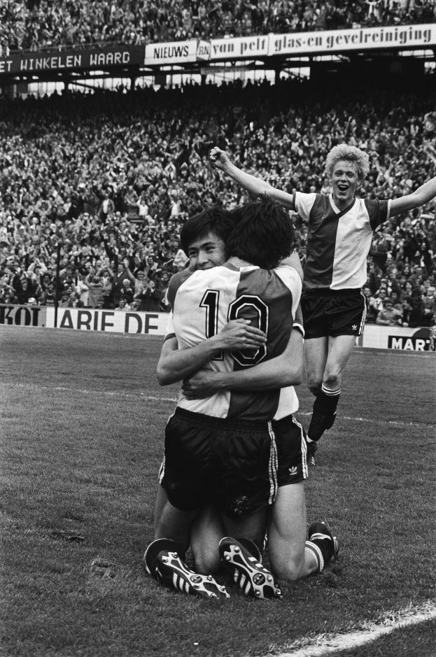 Drei Fußballspieler umarmen sich auf dem Boden nach einem Tor, mit jubelnder Menge und schwenkenden Fahnen im Hintergrund eines Schwarz-Weiß-Foto eines Stadions.