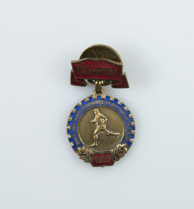 Eine Goldmedaille mit rot-blauem Band, darauf eine laufende Figur und der Text 'Tsempion 1955', vor einem weißen Hintergrund.