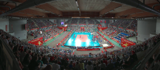 Eine große Halle voller Zuschauer bei einem Volleyballspiel mit Bannern, Bildschirmen, Deckenbeleuchtung und Geländern unten.