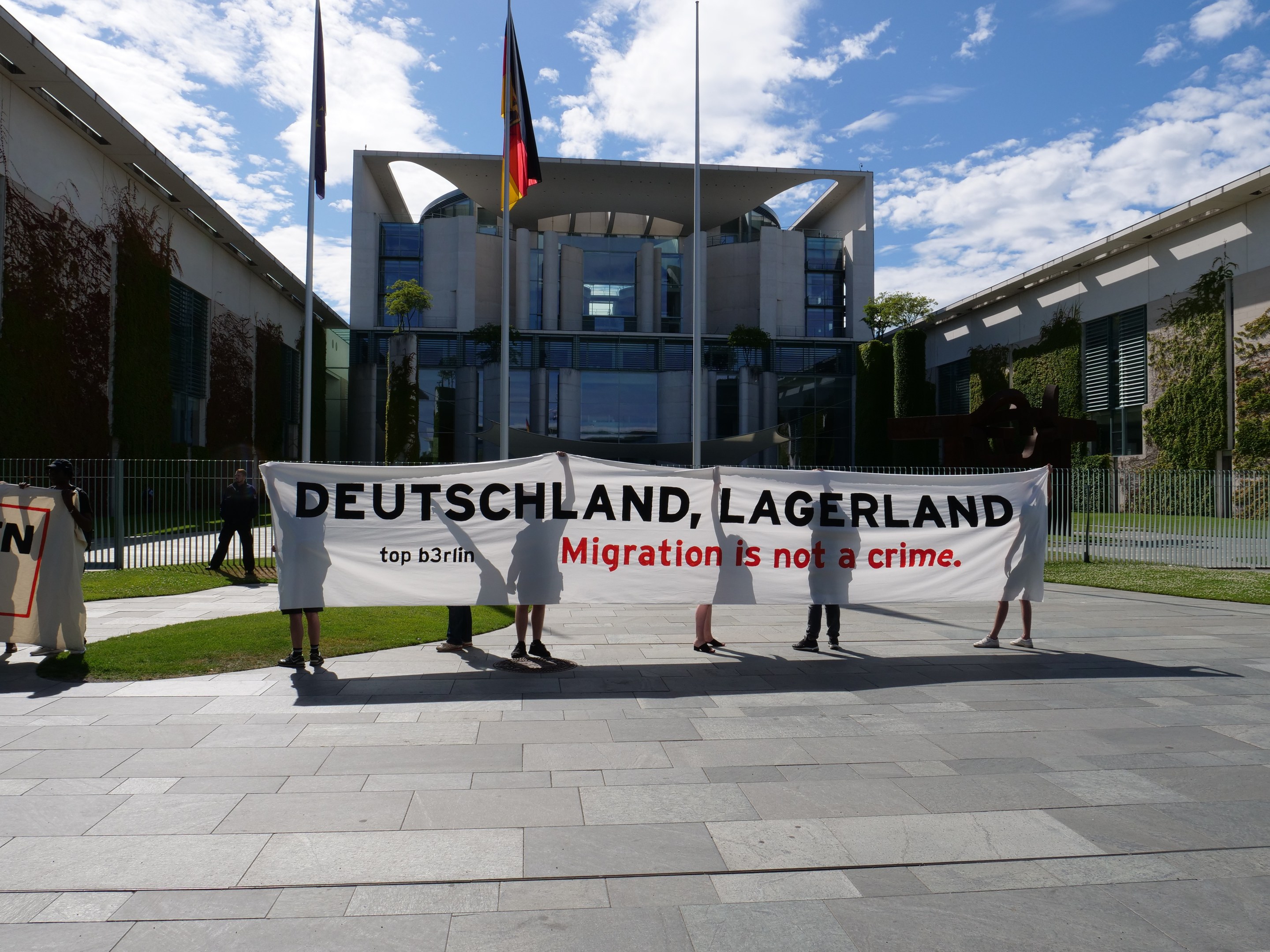 Gruppe von Menschen mit einem Banner "Deutschland, Lagerland Migration ist kein Verbrechen" vor einem Gebäude mit Fenstern, umgeben von Gras, einem Metallzaun, Pflanzen, Bäumen und Fahnenmasten gegen einen bewölkten Himmel.
