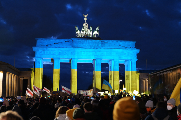 A crowd stands before the Brandenburg Gate in Berlin, holding flags and signs, with a banner on the right displaying protest-related text.