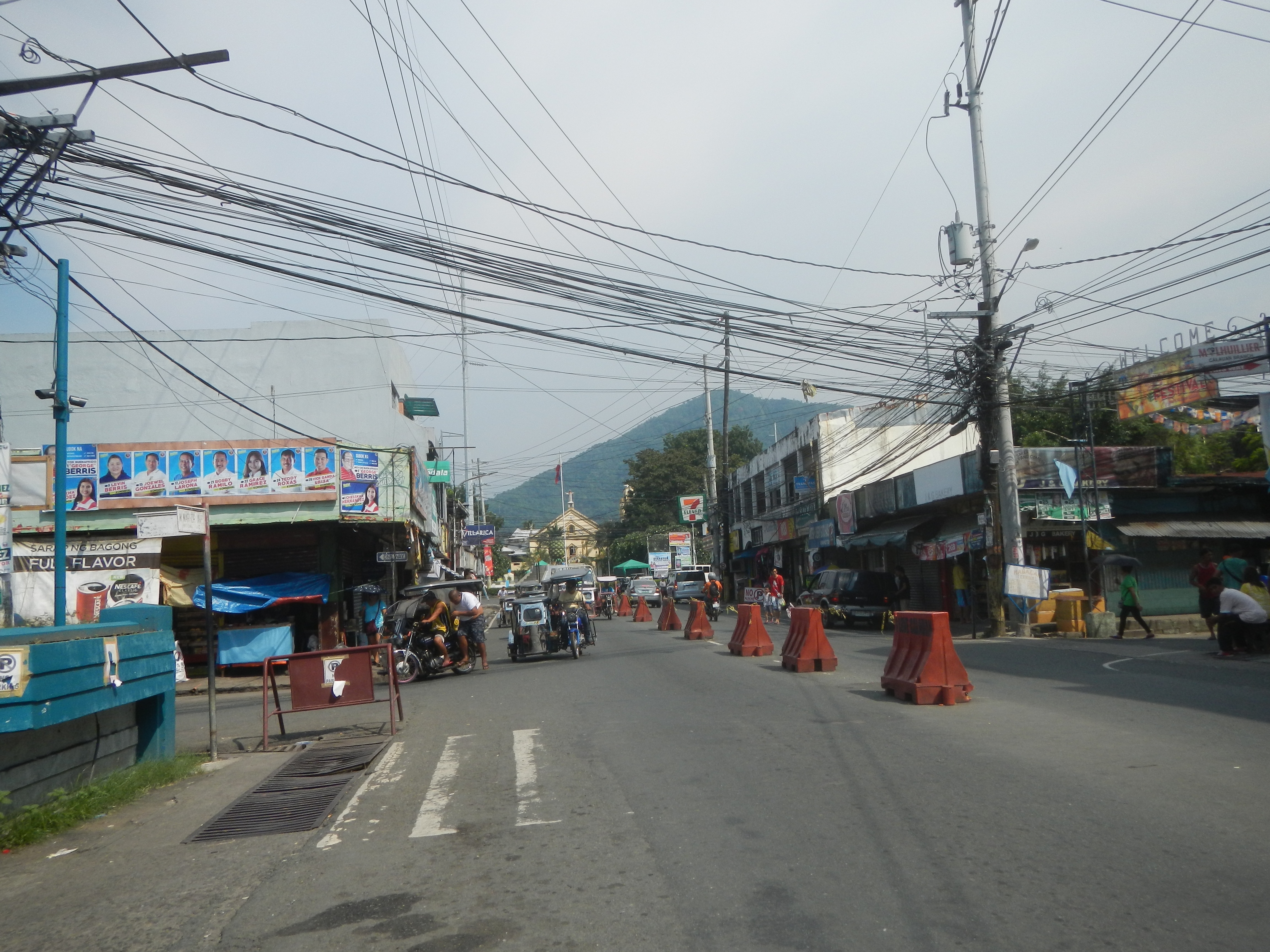 Eine Straßenszene in der Stadt mit Strommasten, Fahrzeugen, Fußgängern, Gebäuden, Werbetafeln, Bäumen, Hügeln und einem klaren blauen Himmel.