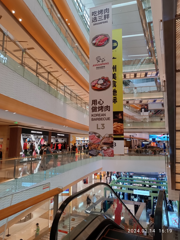 Ein Innenblick in ein Einkaufszentrum mit einer Rolltreppe in der Mitte, Menschen, die umhergehen und Handtaschen tragen, Schilder mit Text an der Decke und Geschäfte im Hintergrund.