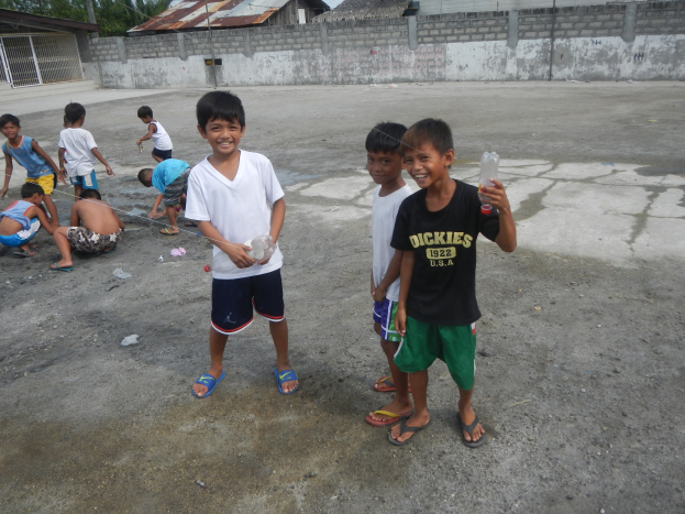 Eine Gruppe junger Jungen steht zusammen in einer Parkanlage, einige halten Flaschen, mit Häusern, Bäumen und Polen im Hintergrund unter einem klaren blauen Himmel.