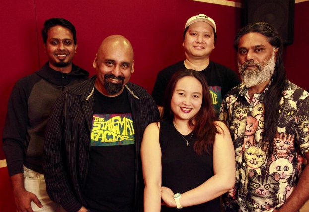 Eine Gruppe von Menschen in legerer Kleidung, die eng zusammenstehen und vor einer roten Wand lächeln, mit einem Lautsprecher im Hintergrund, der darauf hindeutet, dass sie sich in einem Aufnahmestudio befinden.