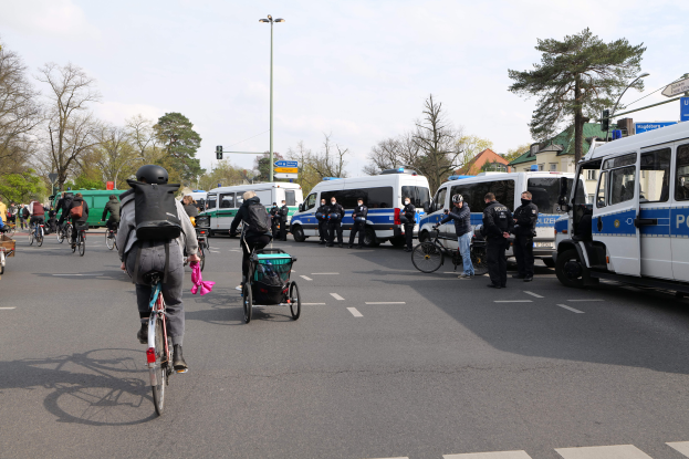 Eine Gruppe von Menschen, die auf Fahrrädern eine Straße entlangfahren, die von Polizeiwagen und anderen Fahrzeugen gesäumt ist, einige tragen Helme und Taschen, andere schieben Kinderwagen, mit Bäumen, Laternen, Verkehrszeichen, Schildern, Gebäuden und einem bewölkten Himmel im Hintergrund.