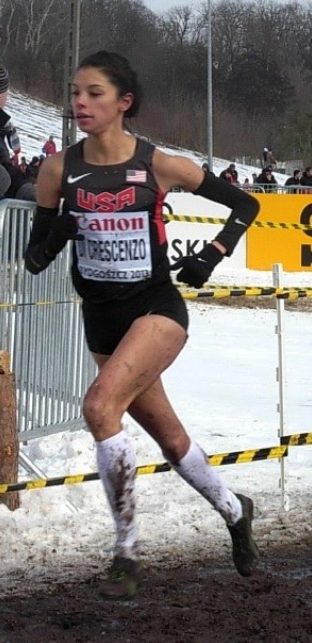 Frau laeuft auf einem schneebedeckten Rennfeld in einem Sportdress, umgeben von einem Metallzaun, Zuschauern, einem Schild, Pfosten, Baeumen und einem bewolkten Himmel.