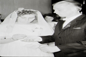 Ein Schwarz-Weiß-Foto einer Braut in einem weißen Kleid und Schleier und eines Bräutigams in einem Anzug, die ihre Eheringe tauschen, mit ein paar Leuten im Hintergrund.