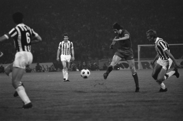 Eine Gruppe von Männern, die Fußball auf einem Feld spielen, mit einem Torpfosten im Hintergrund und Zuschauern, die von der Seitenlinie aus zusehen, dargestellt in Schwarz-Weiß.