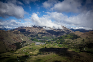 Panoramischer Blick von der Spitze eines Bergs in Queenstown, Neuseeland, mit saftig grünem Gras, verstreuten Bäumen und einer gewundenen Straße unter einem Himmel voller weißer, flauschiger Wolken.
