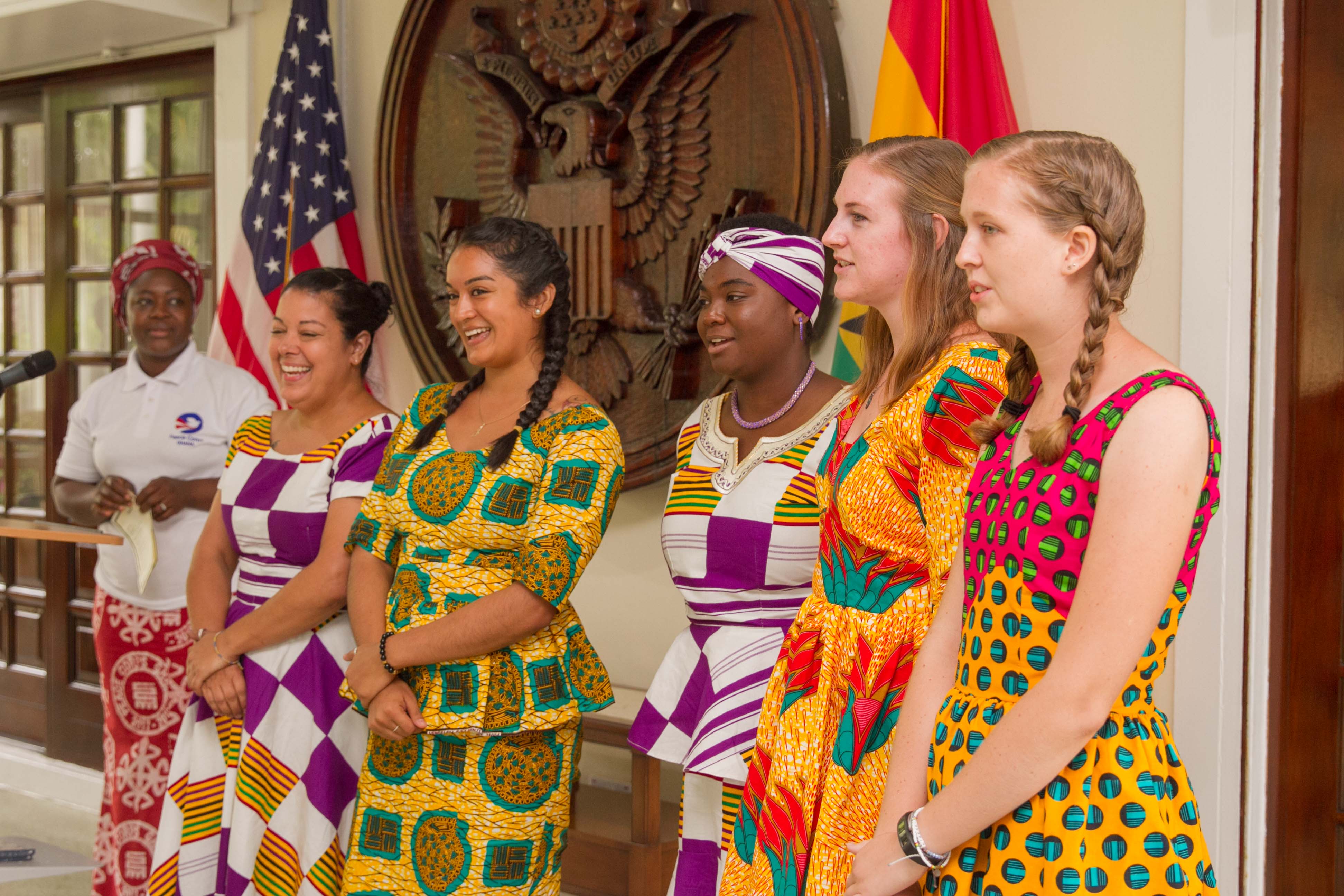 Eine Gruppe von Frauen steht zusammen vor Flaggen, wobei eine Frau neben einem Lautsprecherständer mit einem Mikrofon und einem Gerät steht und ein Emblem und eine Tür an der Rückwand zu sehen sind.