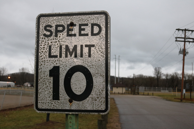 Geschwindigkeitsbegrenzungsschild am Straßenrand mit umliegender Wiese, Strommasten, einem Zaun, Häusern, Bäumen und einem bewölkten Himmel im Hintergrund.