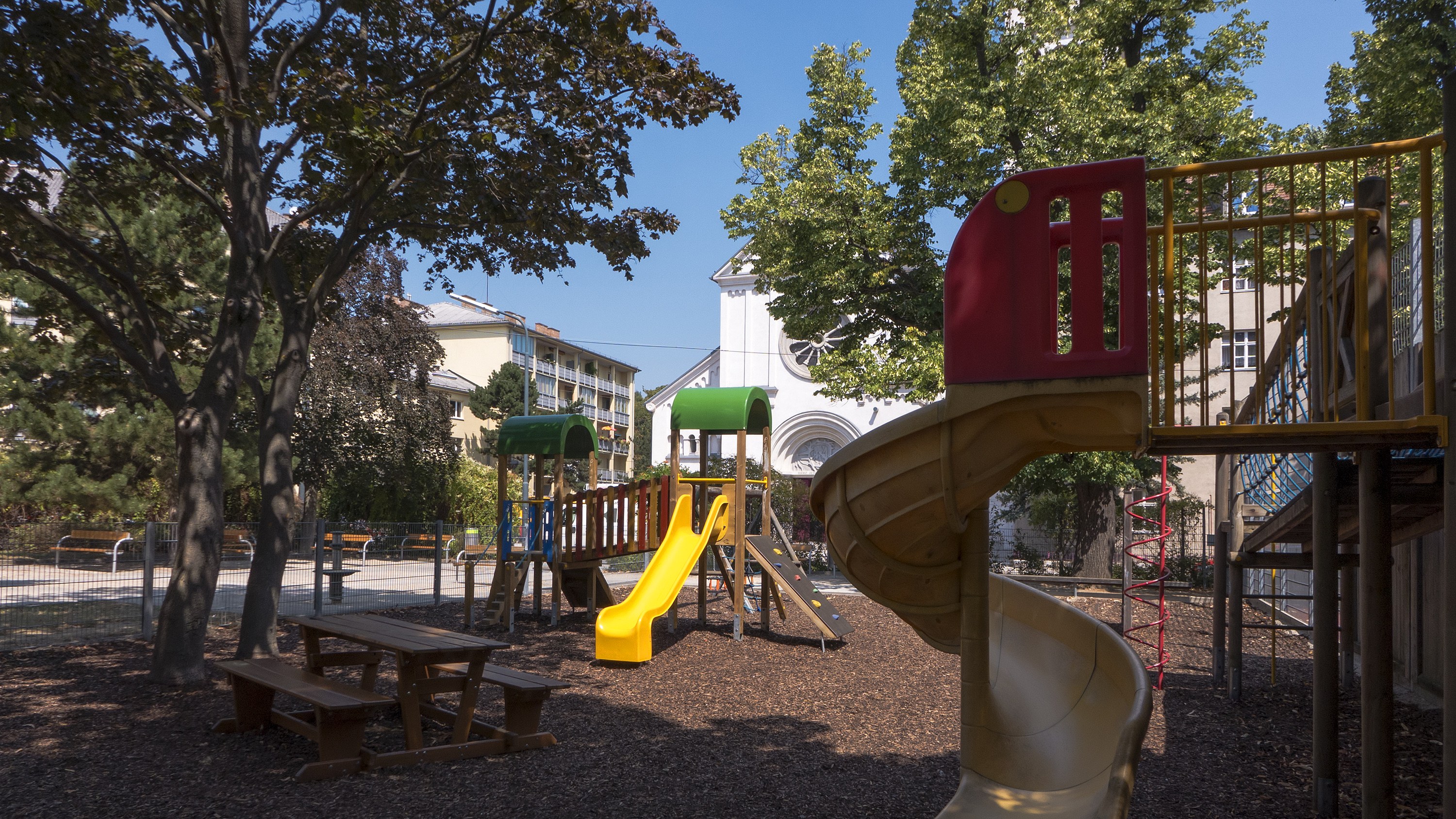Ein Spielplatz mit einer gelben und roten Rutsche, umgeben von Bäumen, Bänken, einem Zaun, Gebäuden und einem klaren blauen Himmel.