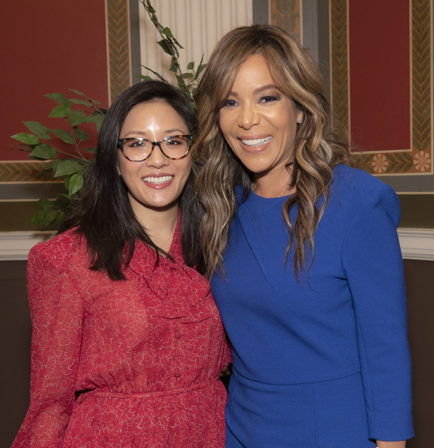 Zwei Frauen, die nebeneinander auf einer Veranstaltung lächeln, eine in einem blauen Kleid und die andere in einem roten Kleid mit Brille, mit einer Pflanze und einem Vorhang im Hintergrund.