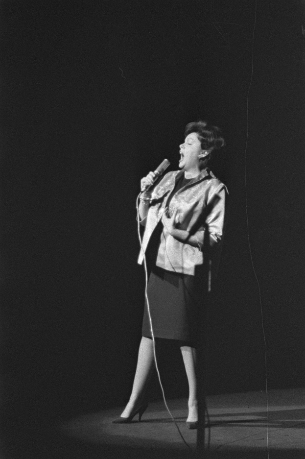 Eine Frau in einem schwarzen und weißen Outfit aus den 1950er-Jahren singt fröhlich in ein Mikrofon auf einer von einem Scheinwerfer beleuchteten Bühne.