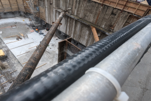 Eine Baustelle mit einem großen Metallrohr in der Mitte, umgeben von Arbeitern, Holzobjekten und diverser Ausrüstung, mit Fahrzeugen und einem Gebäude im Hintergrund.