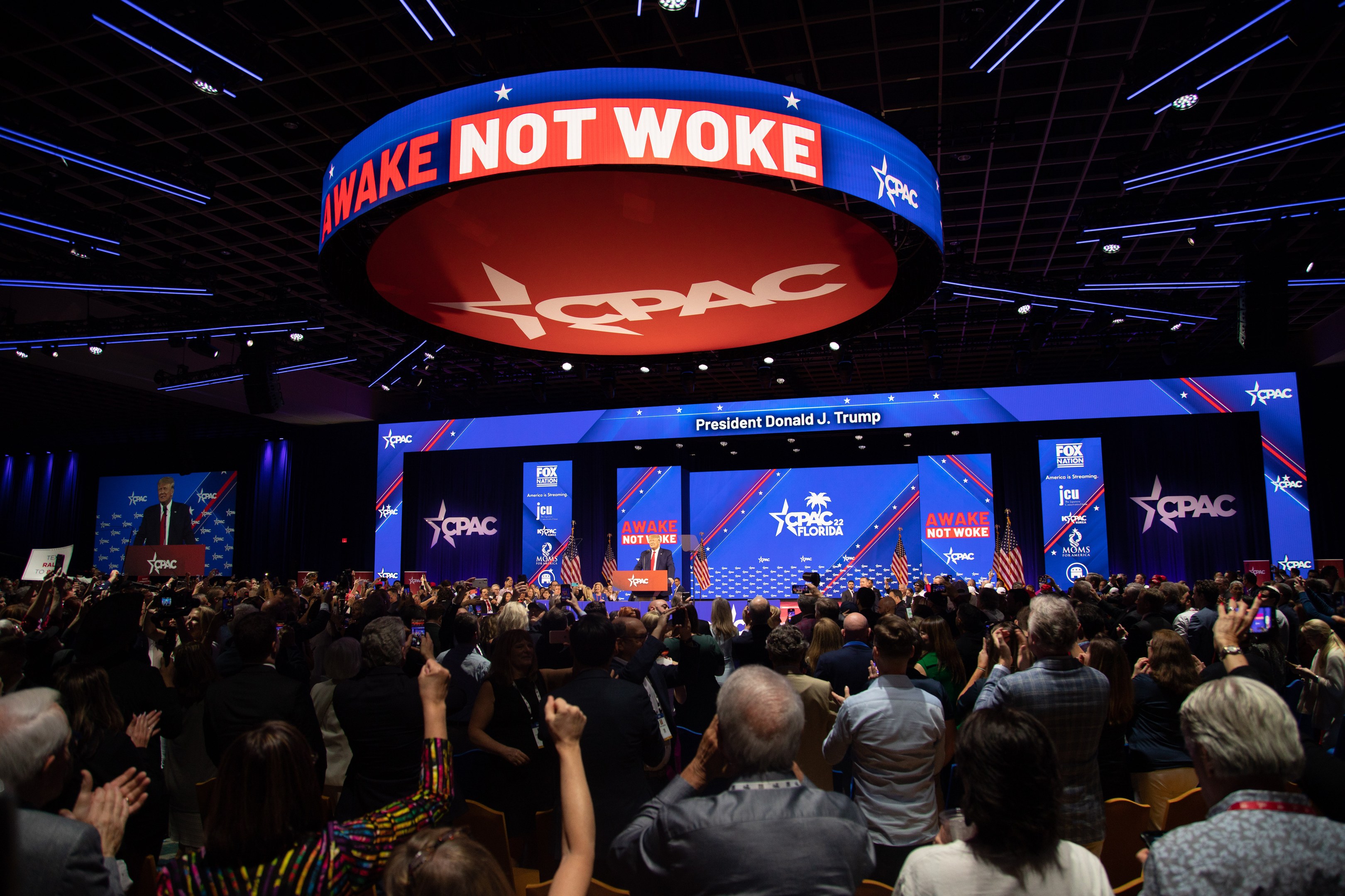 Eine große Menschenmenge sitzt vor einer Bühne mit einem "Wake Not Woke"-Schild auf der Republican National Convention, wo mehrere Personen auf der Bühne unter Deckenlampen und Bildschirmen stehen.