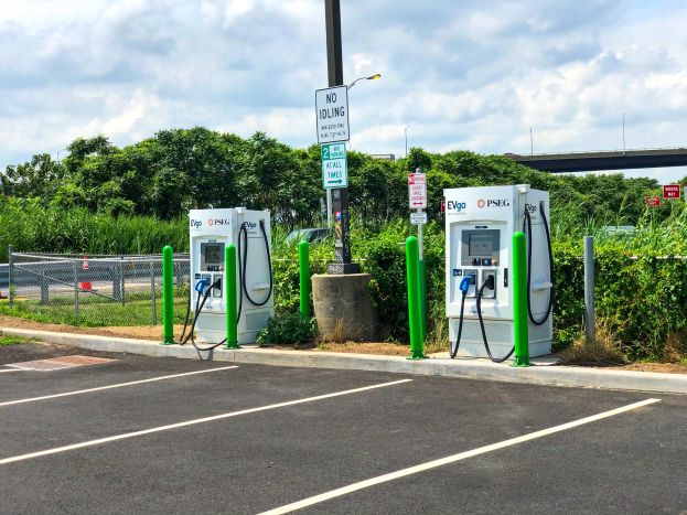 Elektrische Ladestation auf einem Parkplatz mit umgebender Infrastruktur, Grünfläche und einer Brücke im Hintergrund an einem bewölkten Himmel.