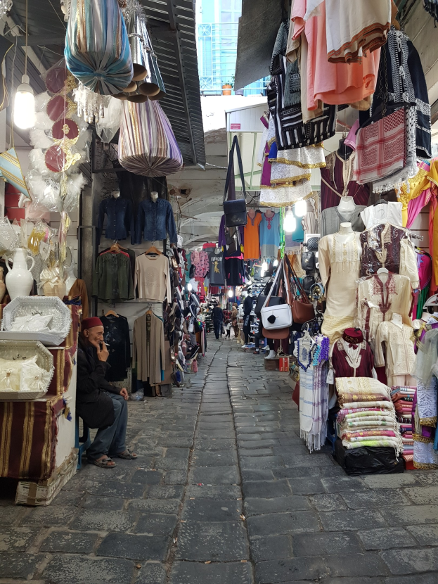 A bustling street market in Jerusalem's old city with stalls selling clothes, bags, lights, and other items, surrounded by people and a building in the background.