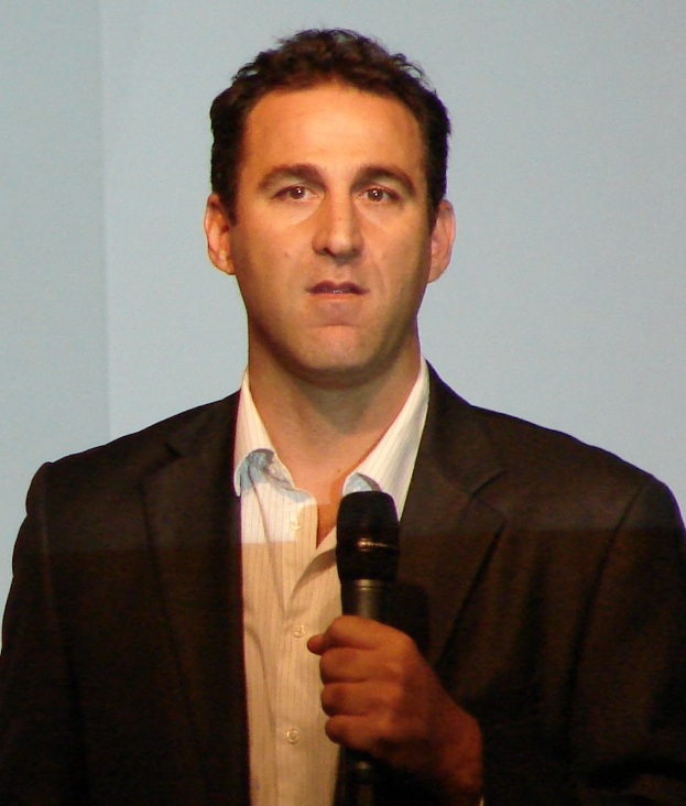 A man in a suit holding a microphone with a serious expression, standing in front of a screen.