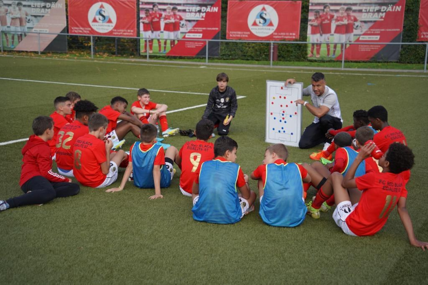 Junge Jungs auf einem Fußballfeld mit einer Tafel vor ihnen, einer einen Fußball haltend, vor einem eingezäunten Bereich mit Bannern und Pflanzen.
