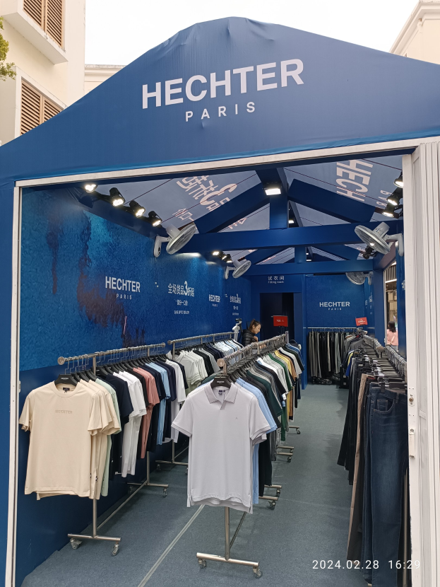 Ein Hechter Paris-Stand auf der Paris Fashion Week, mit Kleidung an Bügeln, einer Person im Stand, Lichtern, einem Gebäude im Hintergrund, einem Baum auf der linken Seite und einem sichtbaren Himmel oben.