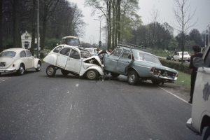 Zwei Autos nach einem Unfall auf der Straße stehend, mit Menschen drumherum, Bäumen, Polen, Gebäuden und einem klaren blauen Himmel im Hintergrund.