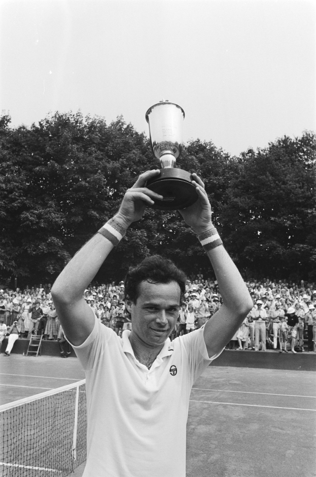 Ein Mann hält einen Pokal auf einem Tennisplatz, umgeben von einem Netz, mit einer Menge und Bäumen im Hintergrund unter einem klaren blauen Himmel.