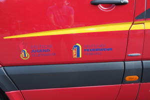 Ein roter Van mit einer gelben Streifen, einem Türgriff und einem Logo mit einem Schild, einem Löwen und einem Lorbeerkranz, sowie der Aufschrift "Deutsche Jugendfeuerwehrverband".