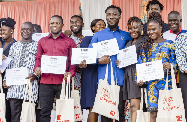 A group of people holding certificates and smiling, with bags at their feet, standing in front of curtains and a wall.