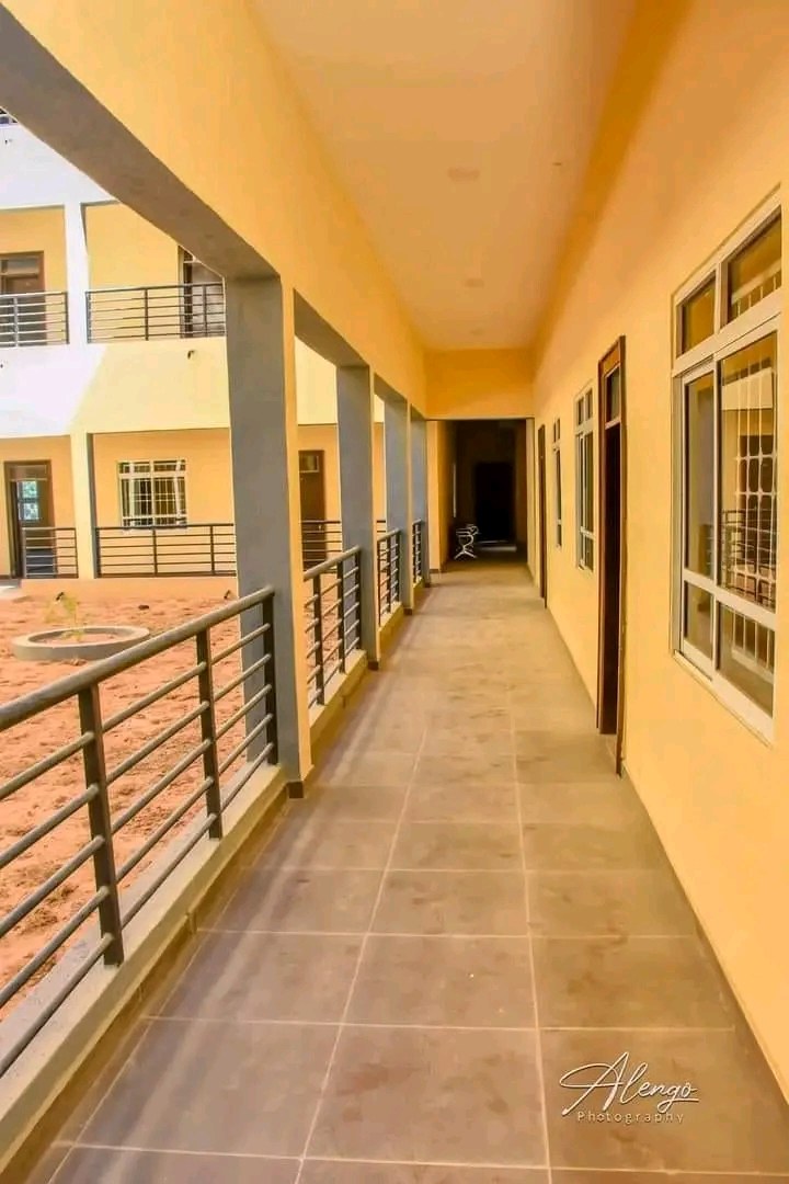 A multi-story building with windows, doors, pillars, and railings, featuring a long tiled hallway and a balcony, set against other buildings and a clear blue sky, with rental text at the bottom.