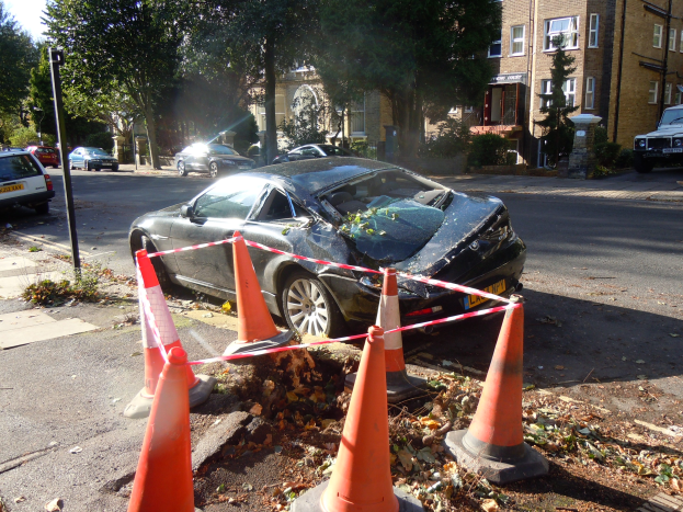 Ein besch├Ądigt auf der Stra├če stehendes Auto mit orangefarbenen Verkehrskegeln drumherum, vor einem Hintergrund aus B├Ąumen, Geb├Ąuden und einem klaren blauen Himmel.