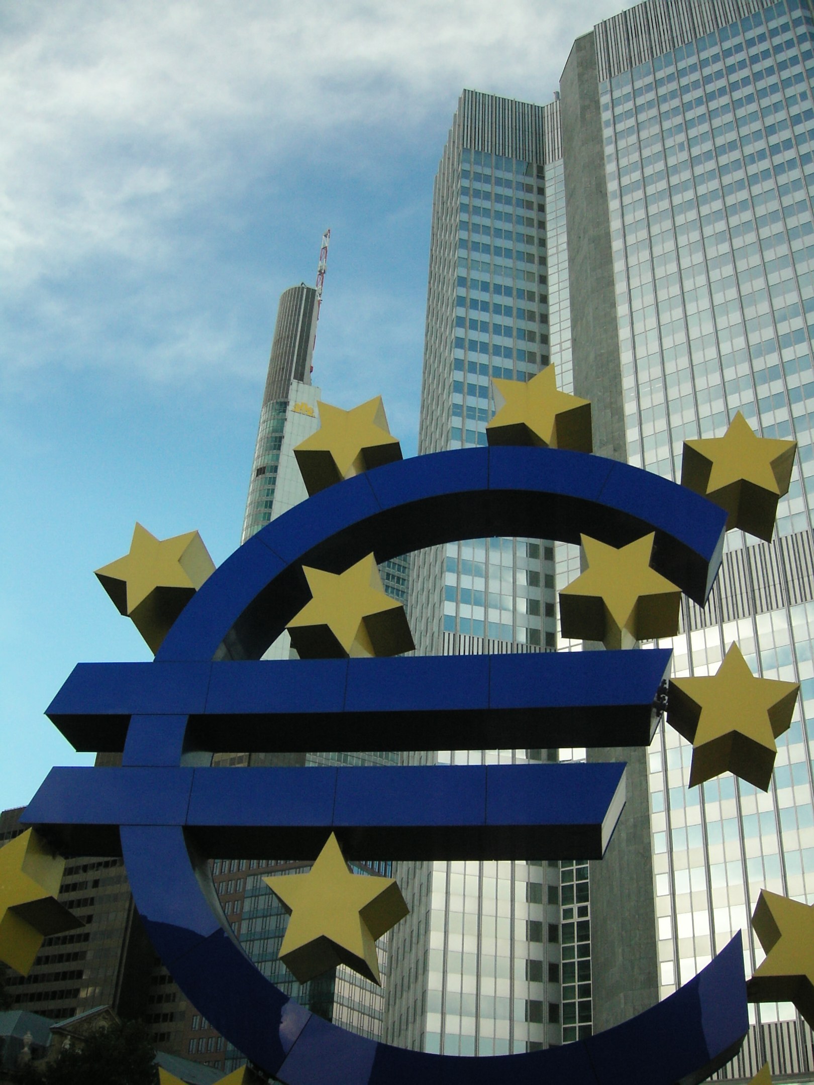 Euro sign in front of the European Central Bank headquarters in Frankfurt, Germany, with the building surrounded by trees under a cloudy sky.