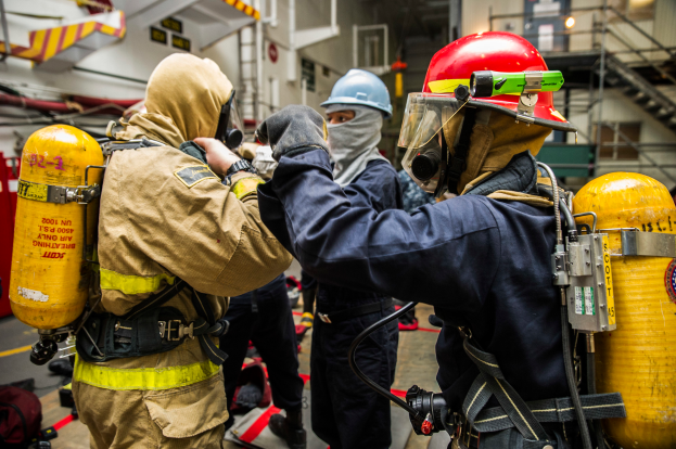 Eine Gruppe von Feuerwehrleuten in Schutzausrüstung, einschließlich Helmen, Handschuhen und Sauerstoffflaschen, steht zusammen mit Taschen auf dem Boden, mit Stufen, Geländern, Lampen und Brettern im Hintergrund.