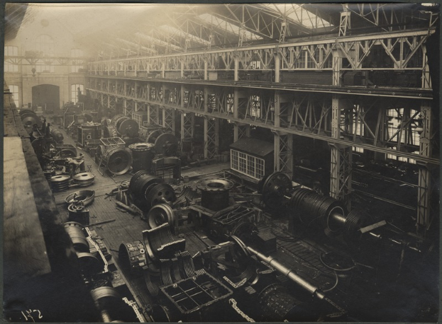 Ein altes Schwarz-Weiß-Foto einer belebten Fabrik mit Maschinen, Stangen und einem Dach, das ein paar Menschen zeigt.