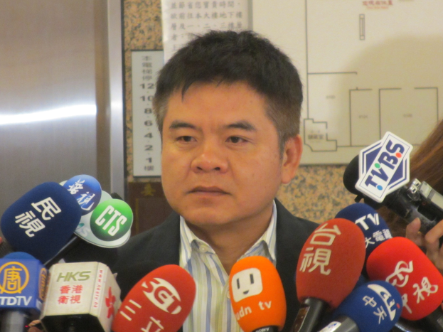 Ein Mann im Anzug spricht vor einer Wand mit einer Tafel zu Reportern. Rechts im Bild hält eine Frau eine Kamera.