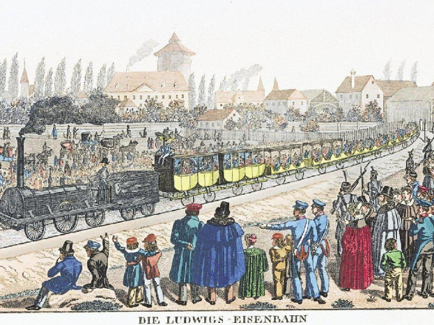 Group of people gathered around a stationary train on tracks, with some passengers inside and others standing outside, against a backdrop of buildings and trees, with text at the bottom reading "Ludwigs-Eisenbahn, the first railway station in Germany."