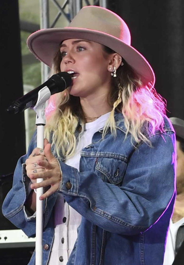 Miley Cyrus performs at the CMA Music Festival in Nashville, Tennessee, singing into a microphone while wearing a hat, with a person in a cap beside her.