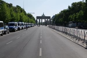 Lange Reihe von Polizeiwagen auf einer Straße vor dem Brandenburger Tor geparkt mit Menschen auf Fahrrädern und Barrieren, Bäume an den Seiten und ein Tor mit Statuen im Hintergrund.