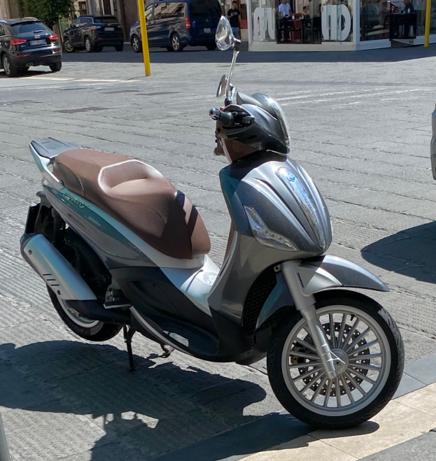 Ein Roller mit Helm auf der Seite einer Straße geparkt, mit Gebäuden, Bäumen, Pfählen, Menschen, Stühlen, Tischen und anderen Gegenständen im Hintergrund.