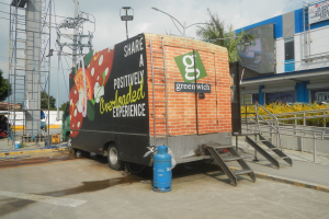 Lkw vor einem Gebäude mit einer Tafel, auf der "Teilen Sie eine positiv überladene Erfahrung" steht, mit Stufen, einem Zylinder am Boden, Geländern, Strommasten mit Drähten, Laternen, Bäumen und einem bewölkten Himmel.